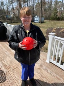 a young girl in a place coat and blue pants stands on a wooden porch holding a red bowling ball