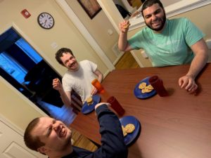 three young men sit at a table with cookies on blue plates and red cups, holding their arms up and smiling