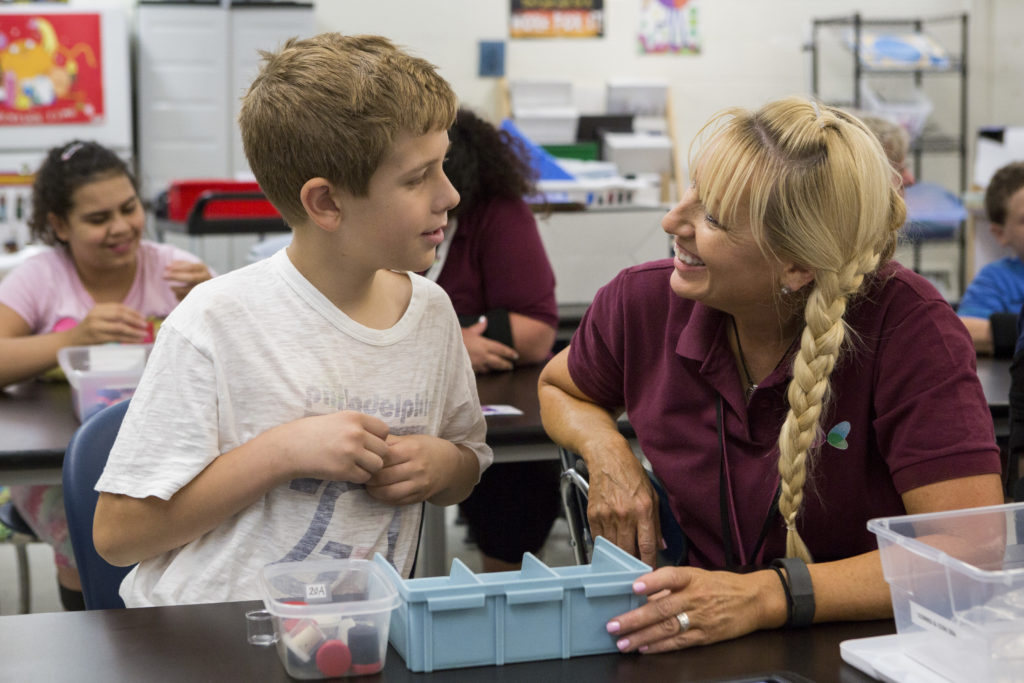 a young boy in a white Philadelphia 76ers shirt sits at a table with a blonde female paraprofessional in a maroon shirt