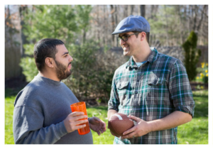 Light-skinned man in a plaid shirt and blue hat holds a football and smiles with a man in a gray sweatshirt