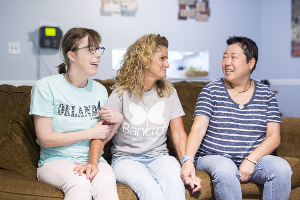 Two females sit on a souch and are smiling at a female direct support professional. They are all holding hands.